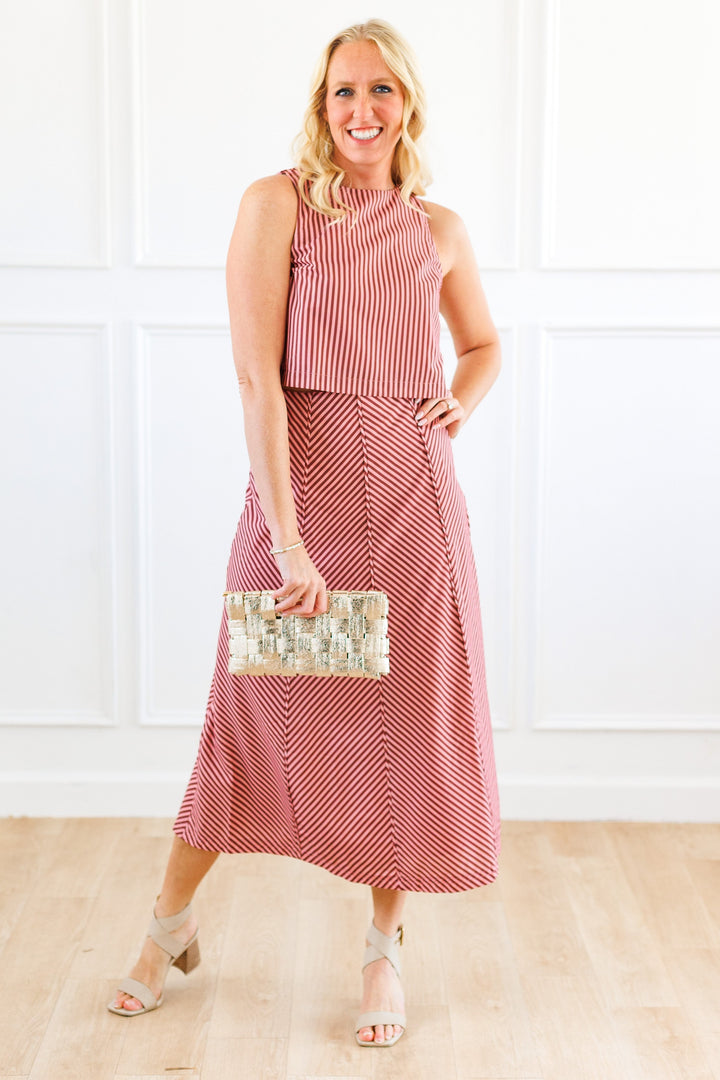 A woman modeling a pink and brown stripe tank and midi skirt set, holding a metallic clutch against a white paneled wall and light wood floor.