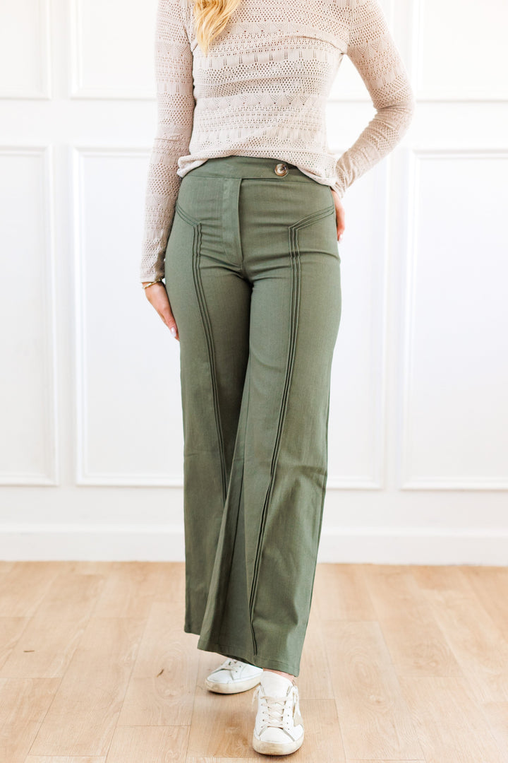 A person wearing olive green wide-leg pants with vertical seam detailing, paired with a cream knit sweater and white sneakers, standing in a bright room with white paneled walls and light wood flooring.