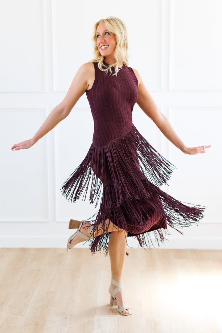 A woman wearing a sleeveless, dark plum-purple fringed dress is captured mid-step in a bright, white-paneled room with light wood floors.