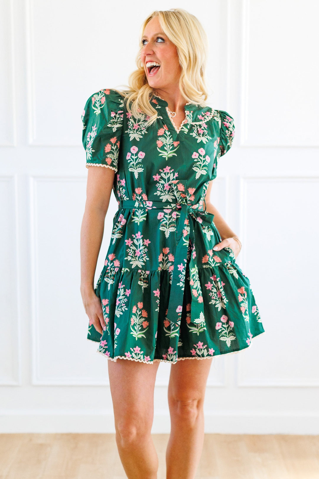 A woman modeling a green floral mini dress with puff sleeves and a tie waist, standing in a bright room with white paneled walls and light wood floors.