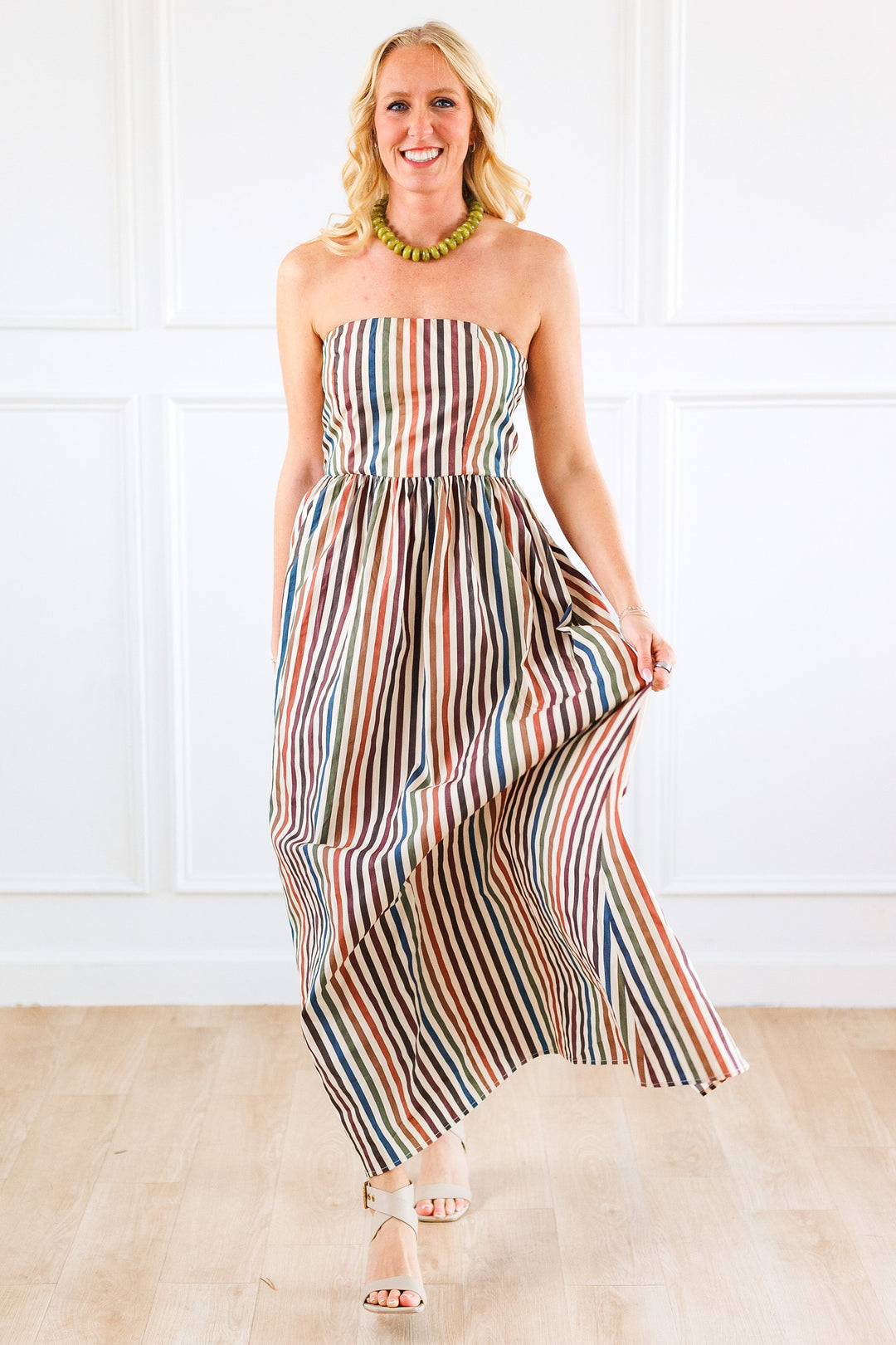 A woman modeling a strapless maxi dress with vertical multicolor stripes in shades of blue, red, orange, and cream, standing on a light wood floor against a white paneled wall.